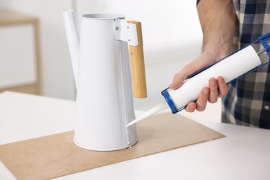 Photo of Man with caulking gun glueing watering can at white table indoors, closeup