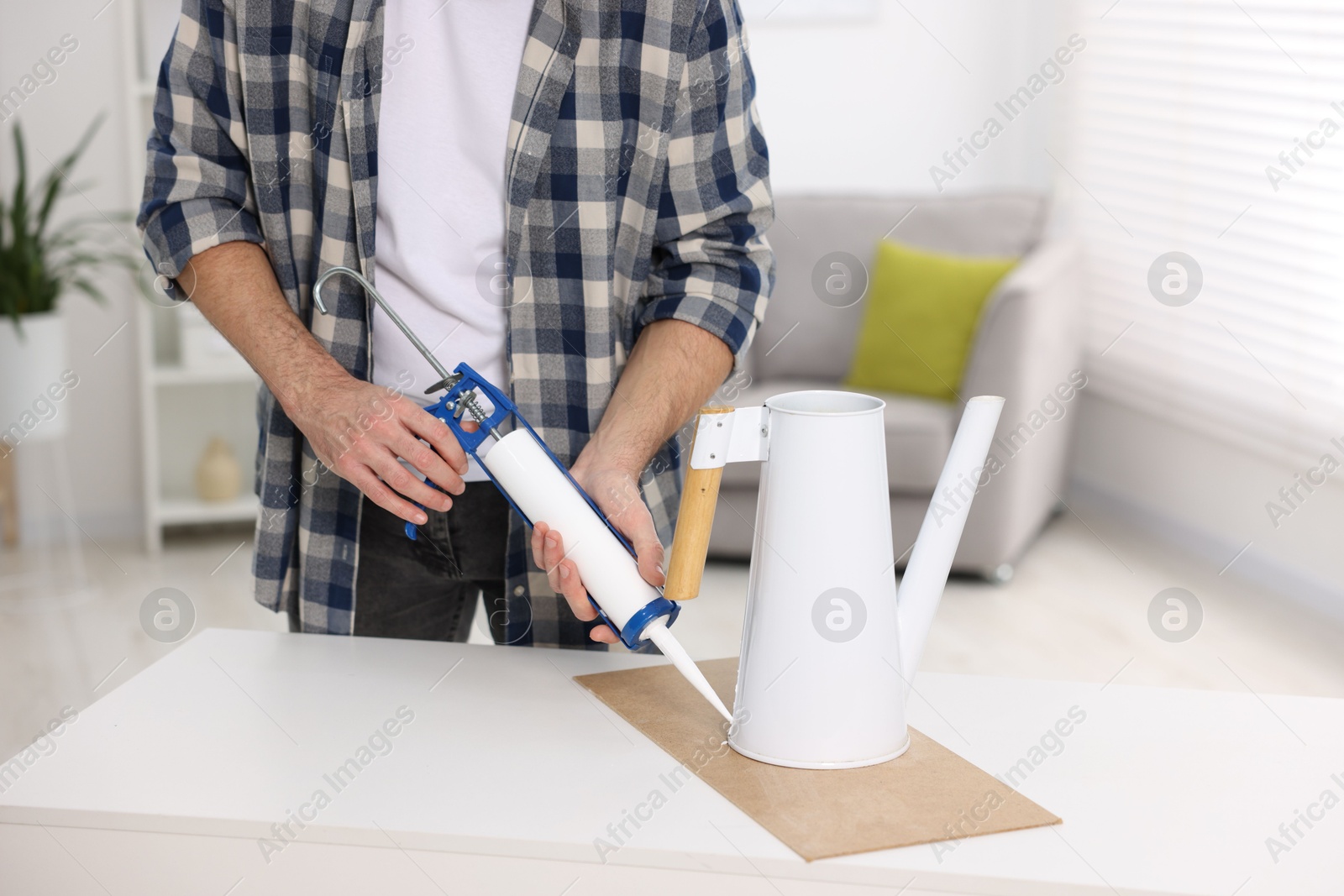 Photo of Man with caulking gun glueing watering can at white table indoors, closeup