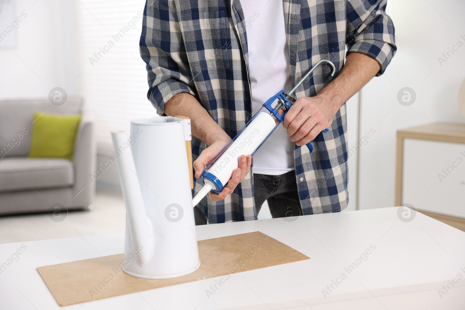 Photo of Man with caulking gun glueing watering can at white table indoors, closeup