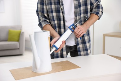 Photo of Man with caulking gun glueing watering can at white table indoors, closeup