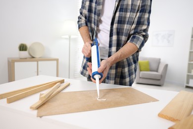 Photo of Man with caulking gun glueing plywood at white table indoors, closeup