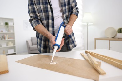 Photo of Man with caulking gun glueing plywood at white table indoors, closeup