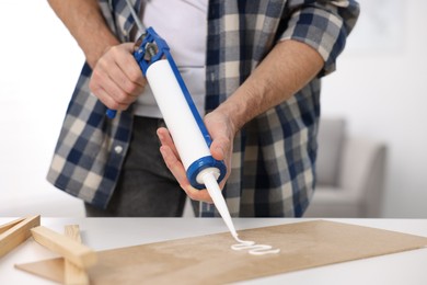 Photo of Man with caulking gun glueing plywood at white table indoors, closeup