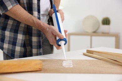 Photo of Man with caulking gun glueing plywood at white table indoors, closeup