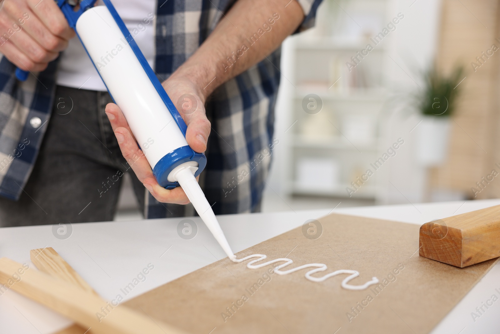 Photo of Man with caulking gun glueing plywood at white table indoors, closeup