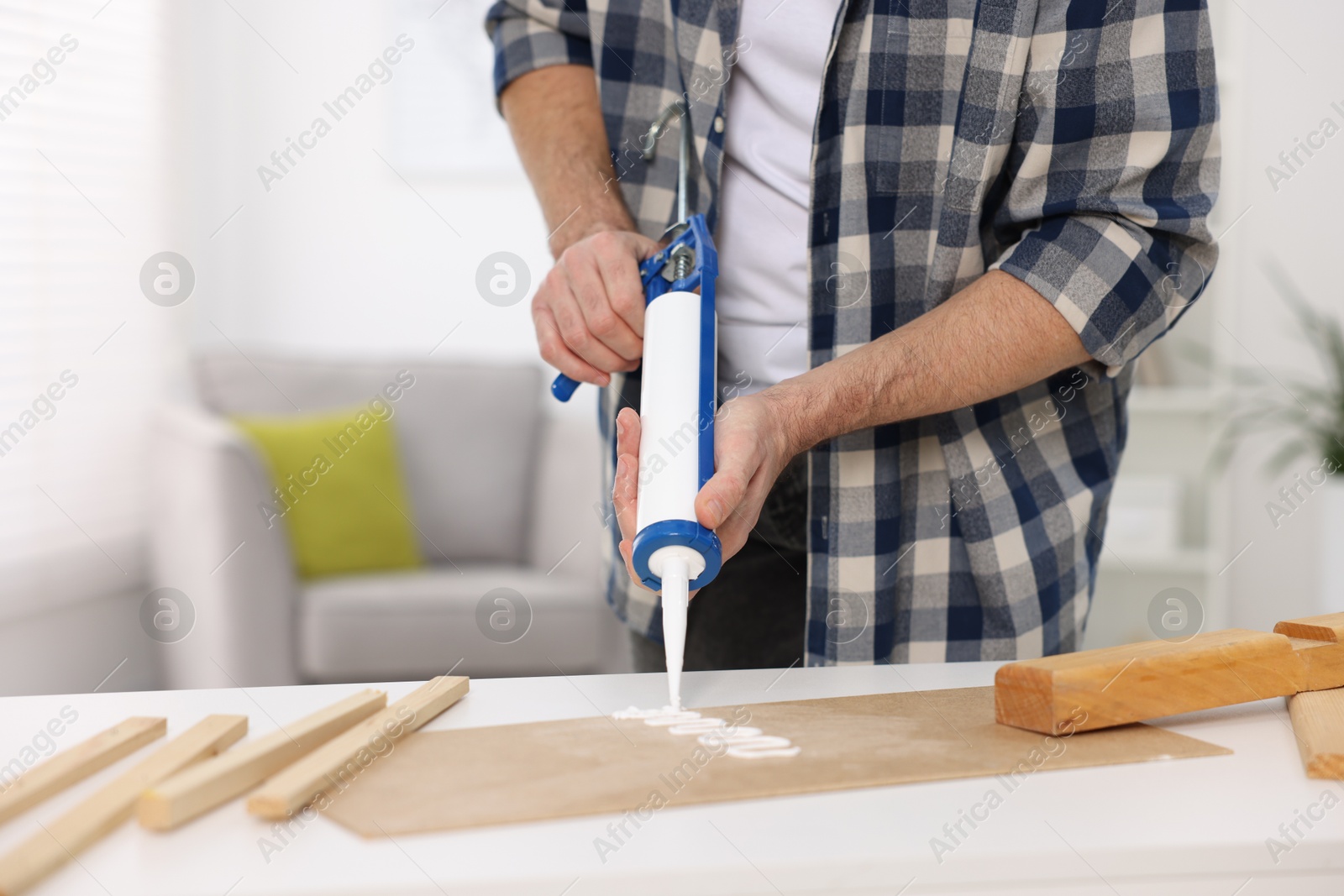 Photo of Man with caulking gun glueing plywood at white table indoors, closeup