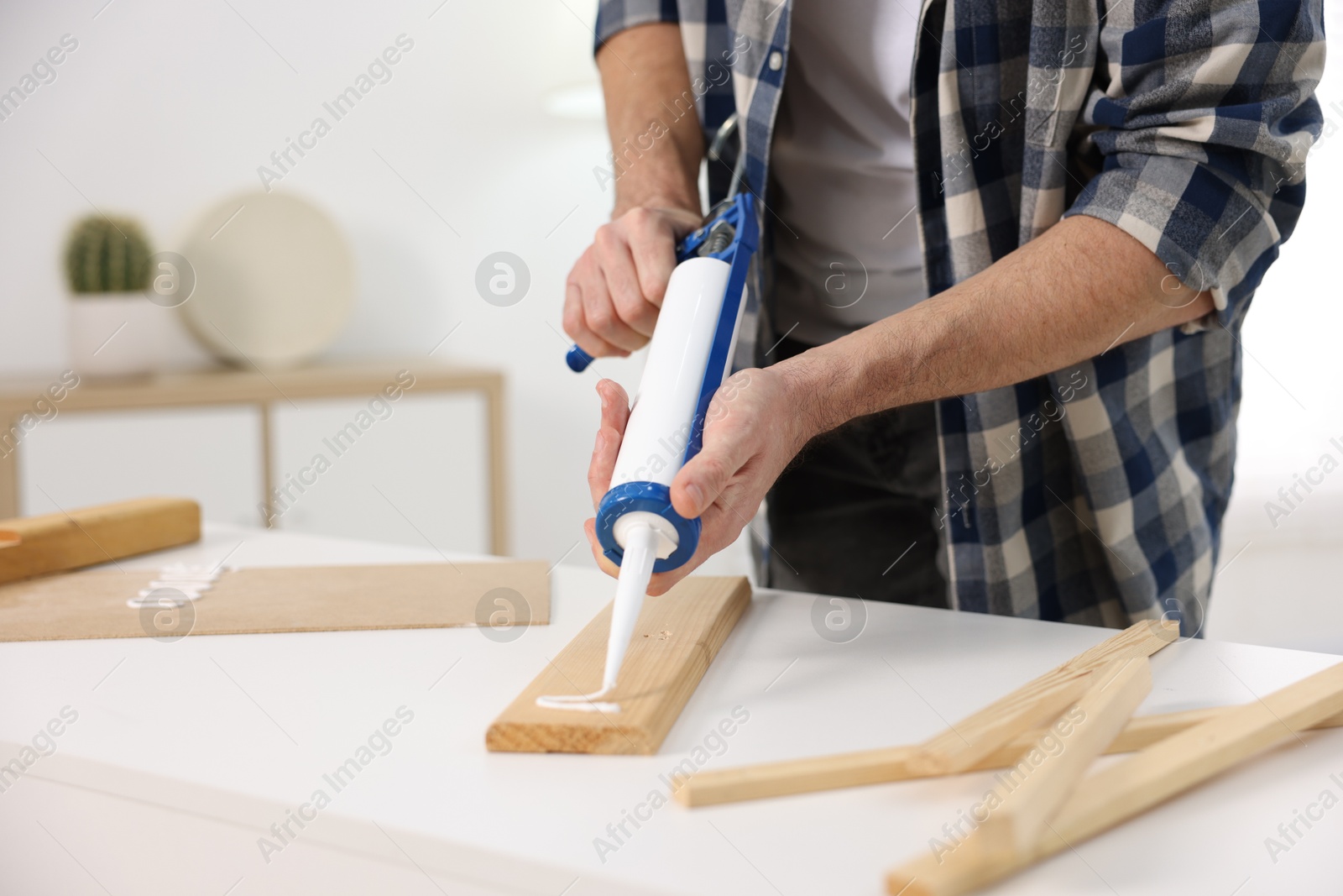 Photo of Man with caulking gun glueing wooden plank indoors, closeup