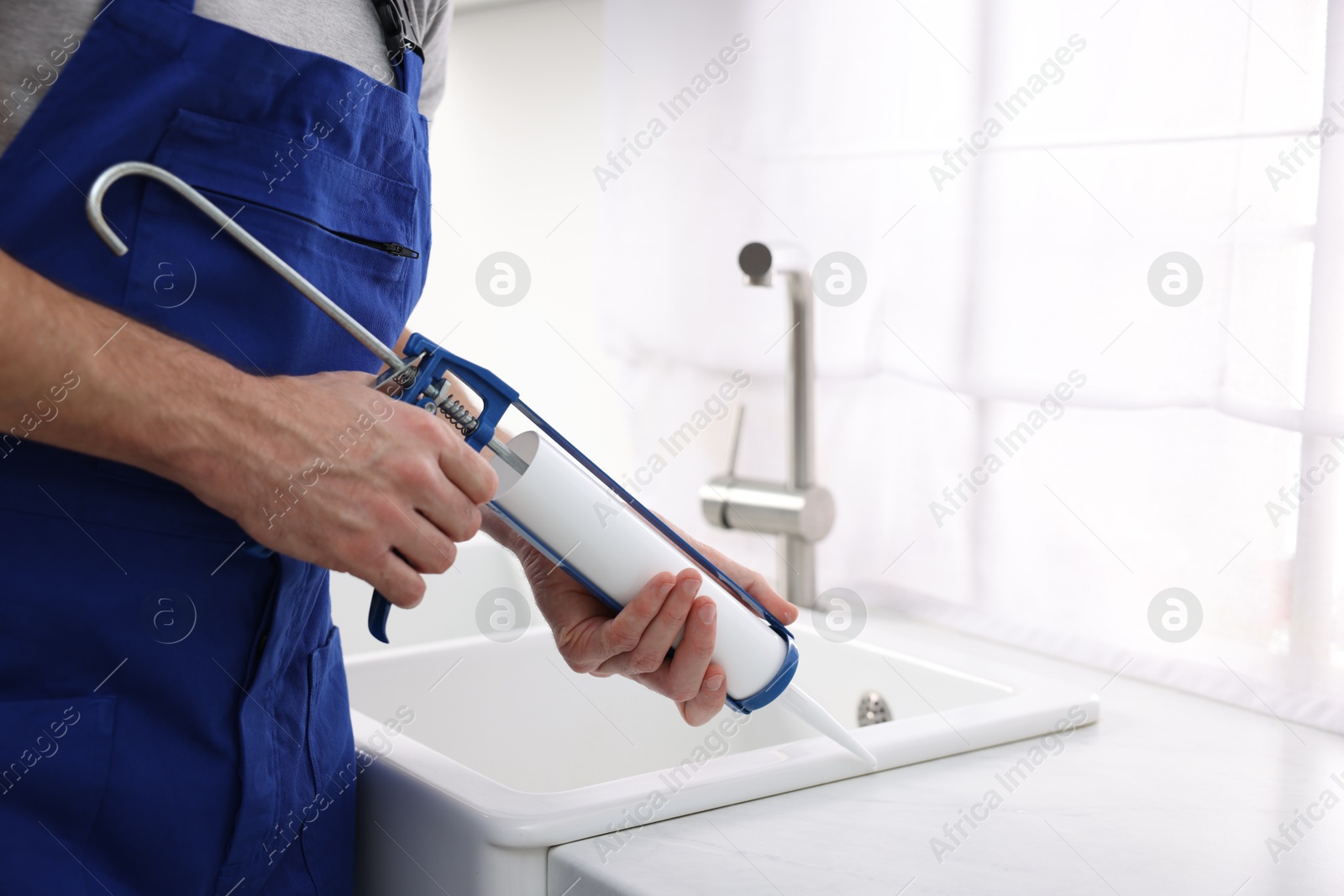 Photo of Worker with caulking gun sealing kitchen sink indoors, closeup