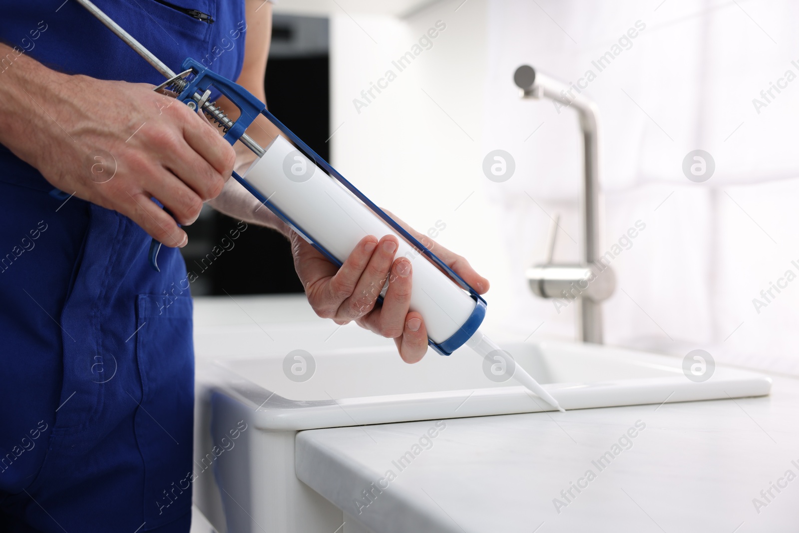 Photo of Worker with caulking gun sealing kitchen sink indoors, closeup