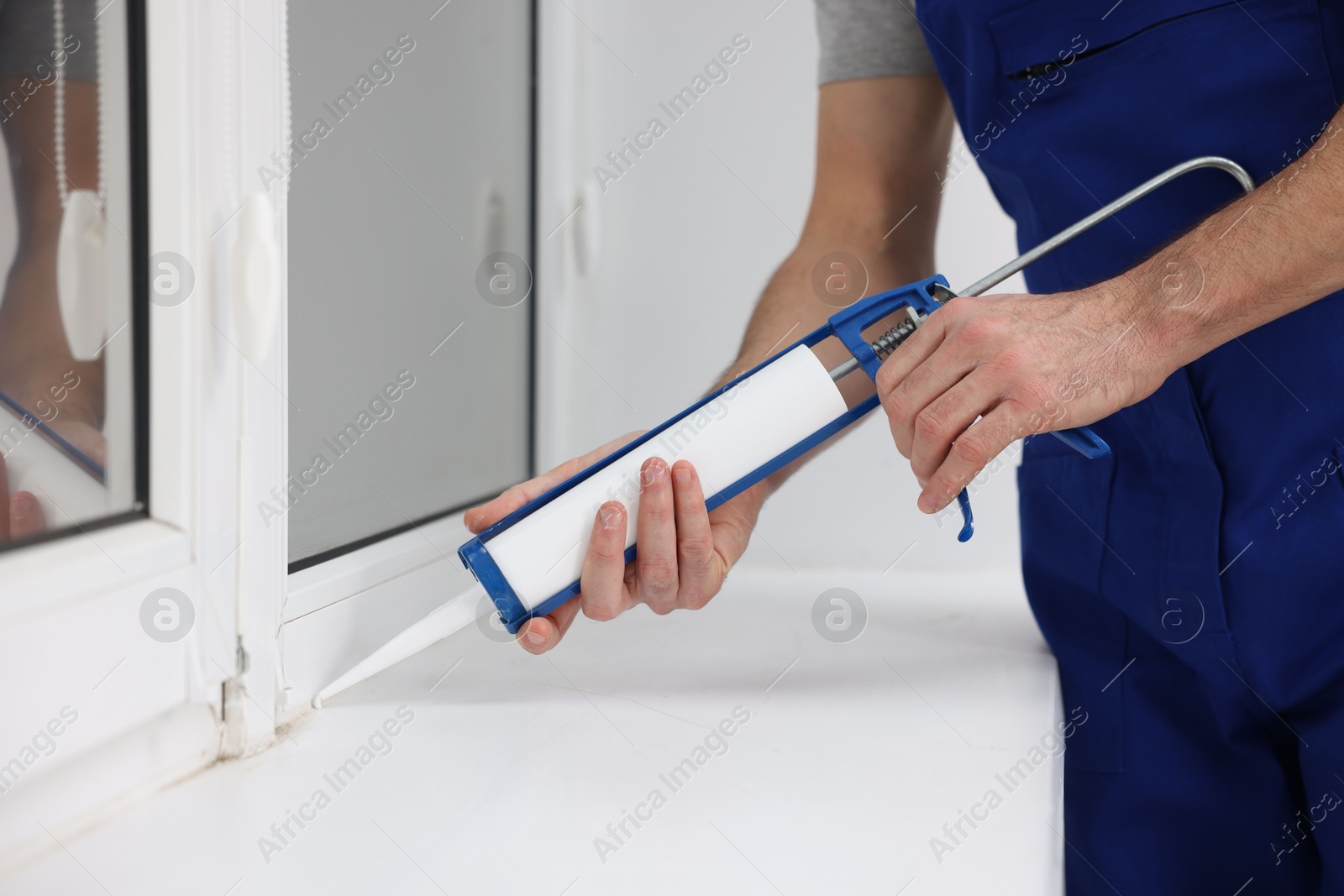 Photo of Worker with caulking gun sealing window indoors, closeup