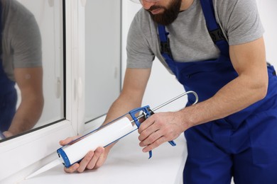 Photo of Worker with caulking gun sealing window indoors, closeup