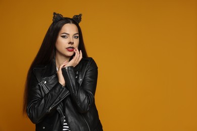 Photo of Woman with cat makeup and ears in leather jacket on orange background. Space for text