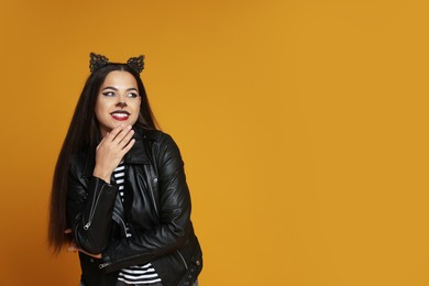 Photo of Woman with cat makeup and ears in leather jacket on orange background. Space for text