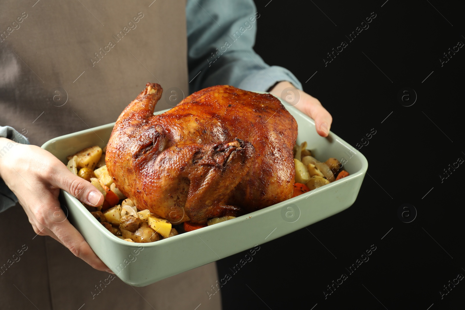 Photo of Woman holding baking tray with delicious baked turkey and vegetables on black background, closeup