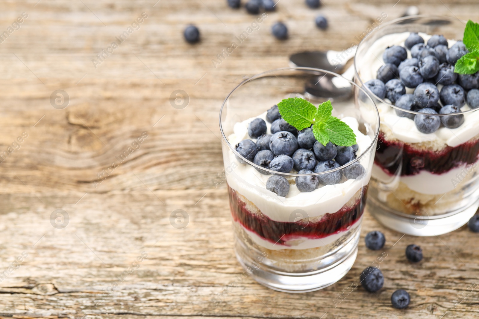 Photo of Tasty trifle dessert. Sponge cake, blueberries, jam and whipped cream in glasses on wooden table, closeup. Space for text