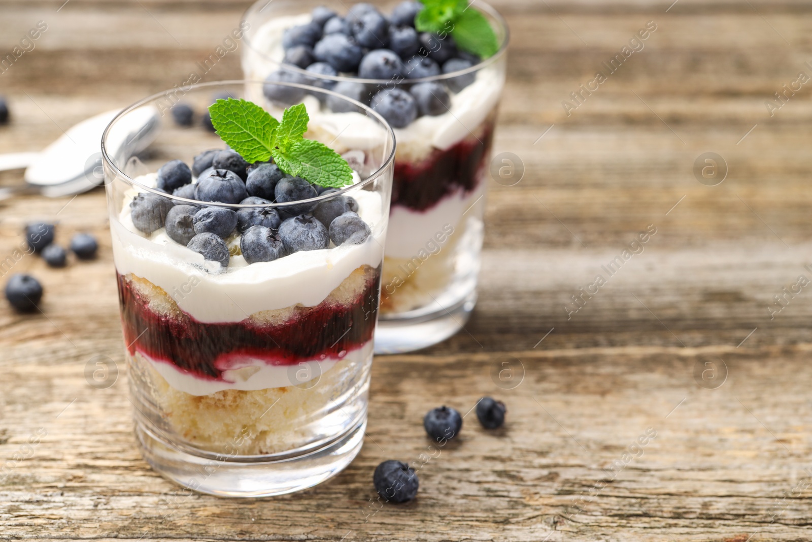 Photo of Tasty trifle dessert. Sponge cake, blueberries, jam and whipped cream in glasses on wooden table, closeup. Space for text