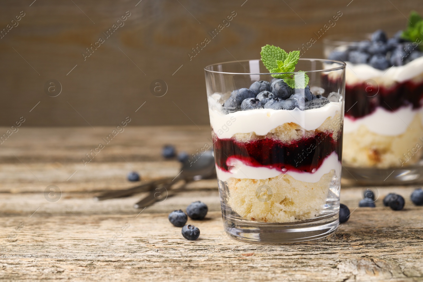 Photo of Tasty trifle dessert. Sponge cake, blueberries, jam and whipped cream in glasses on wooden table, closeup. Space for text