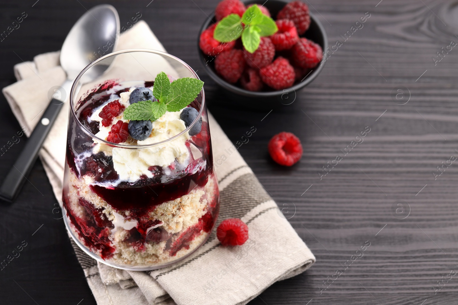 Photo of Tasty trifle dessert. Berries, sponge cake and whipped cream in glass on black wooden table, space for text