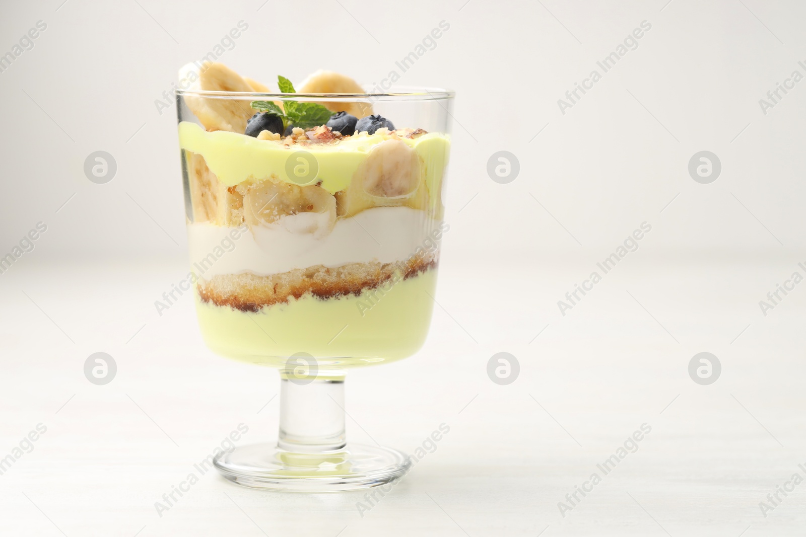 Photo of Tasty trifle dessert. Banana, blueberries, sponge cake and whipped cream in glass on white wooden table, space for text