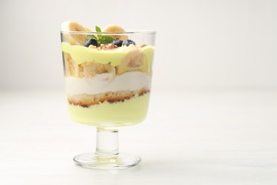 Photo of Tasty trifle dessert. Banana, blueberries, sponge cake and whipped cream in glass on white wooden table, space for text