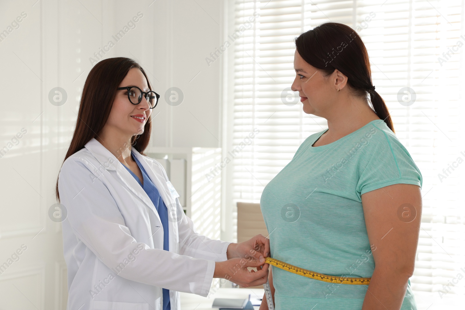 Photo of Nutritionist measuring overweight woman's waist with tape in hospital