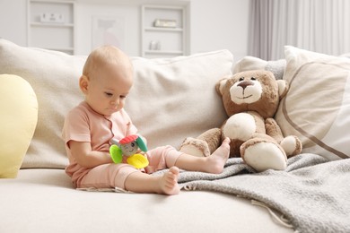 Photo of Cute little baby with rattle and teddy bear on sofa indoors