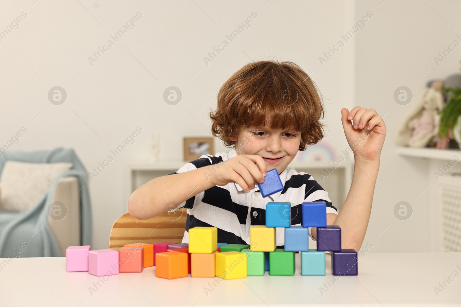 Photo of Little boy stacking colorful cubes at white table indoors