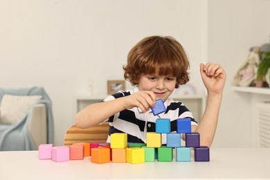 Little boy stacking colorful cubes at white table indoors