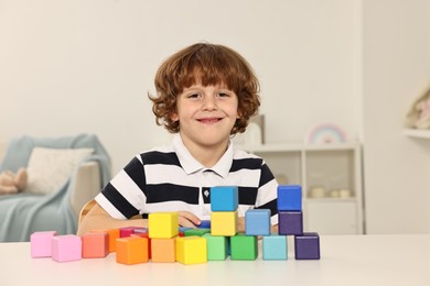 Little boy stacking colorful cubes at white table indoors