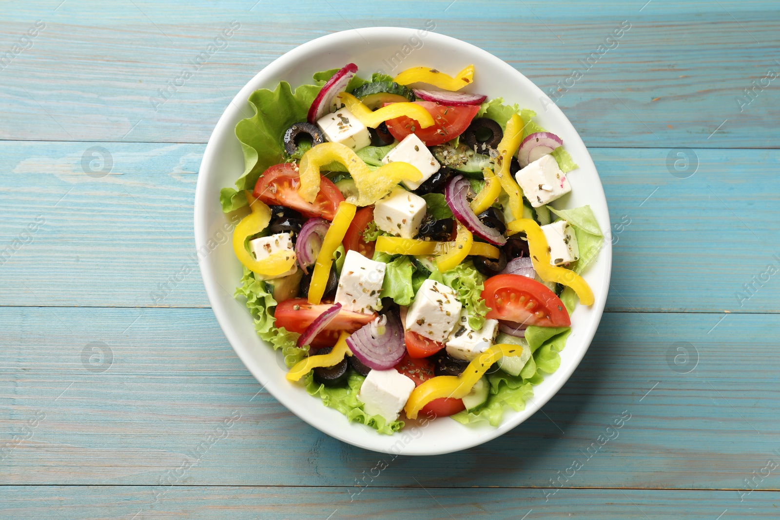 Photo of Delicious fresh Greek salad in bowl on light blue wooden table, top view