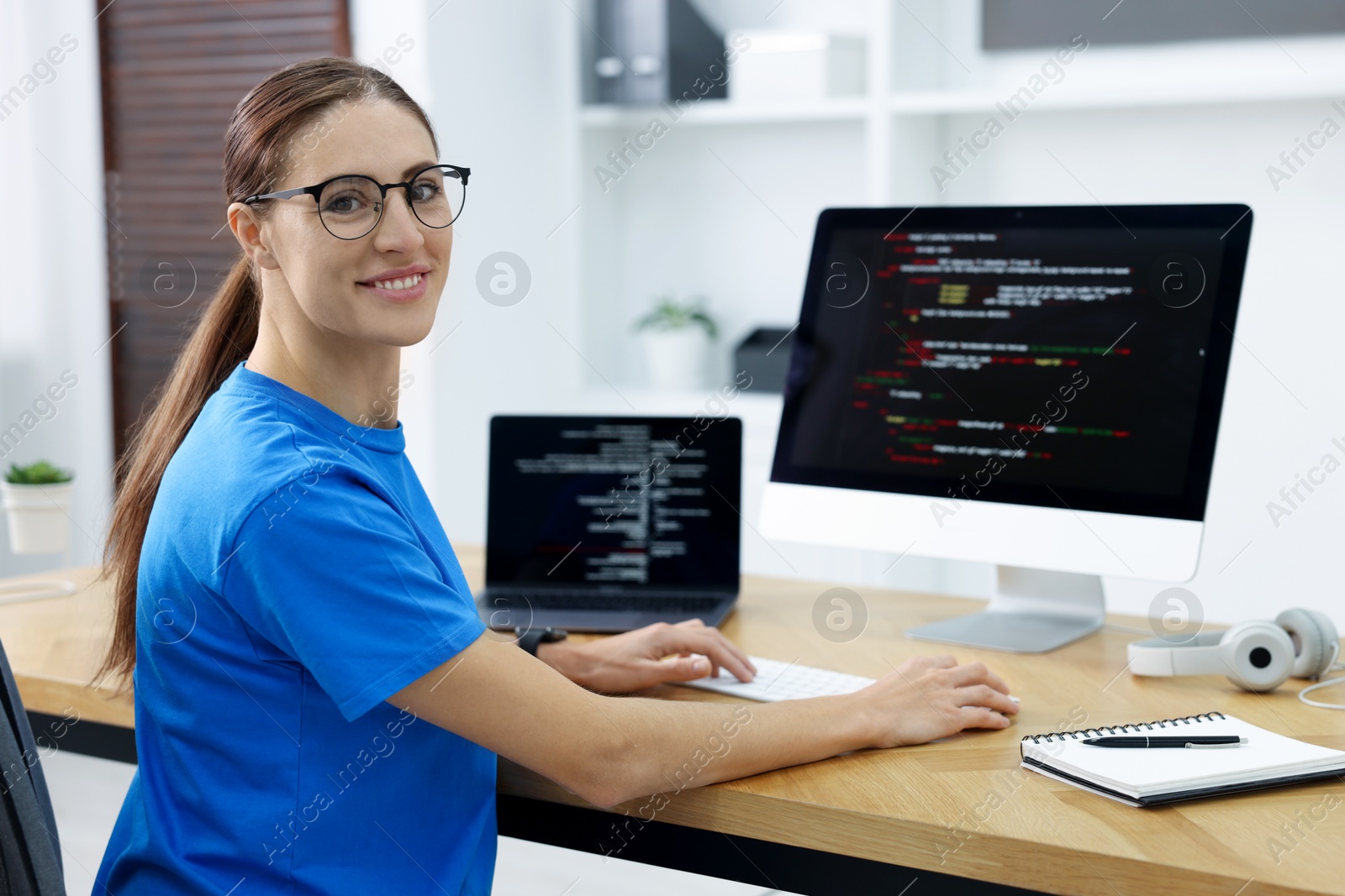 Photo of Programmer working on laptop and computer at wooden desk indoors