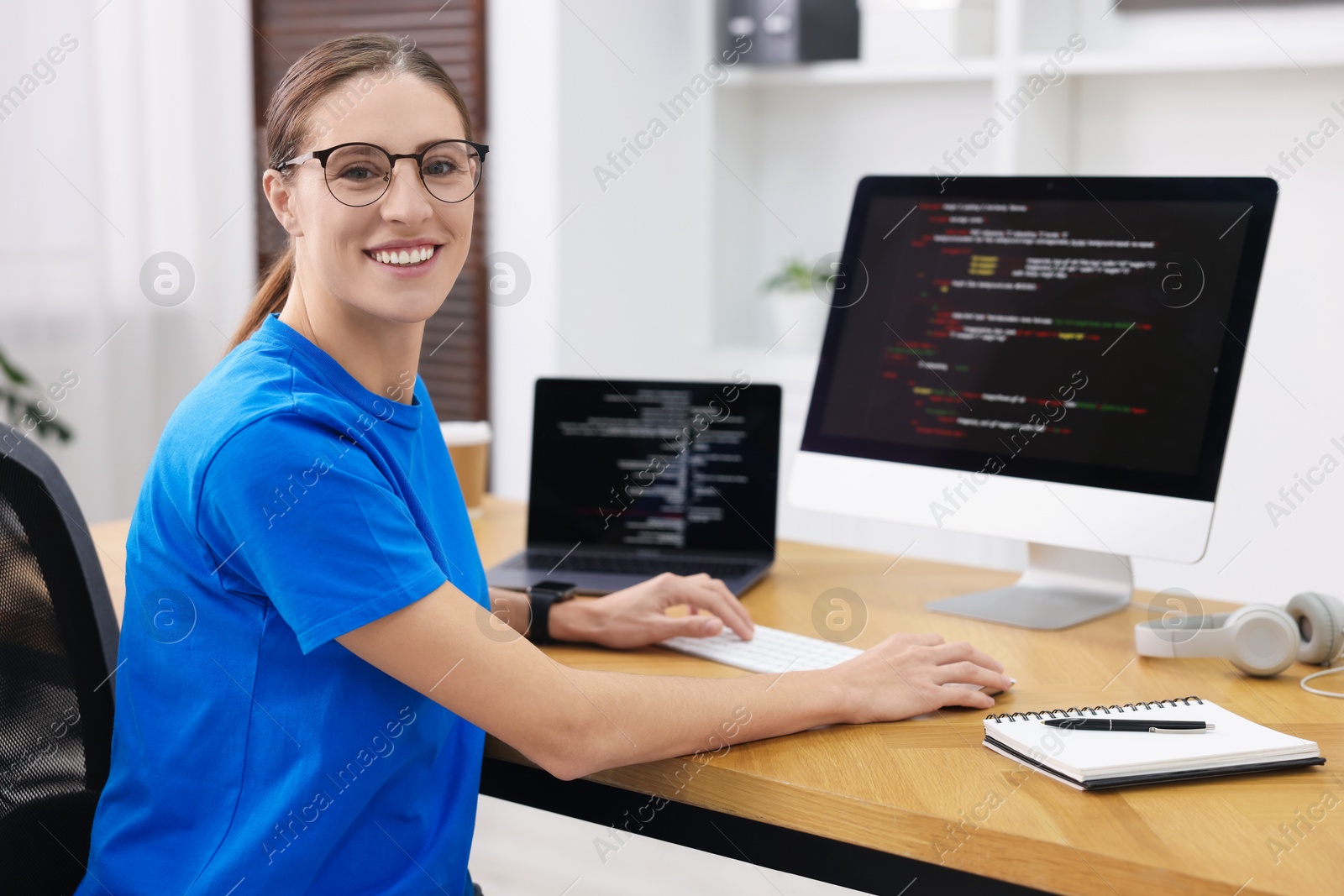 Photo of Programmer working on laptop and computer at wooden desk indoors