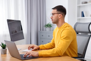 Photo of Programmer working on laptop and computer at wooden desk indoors