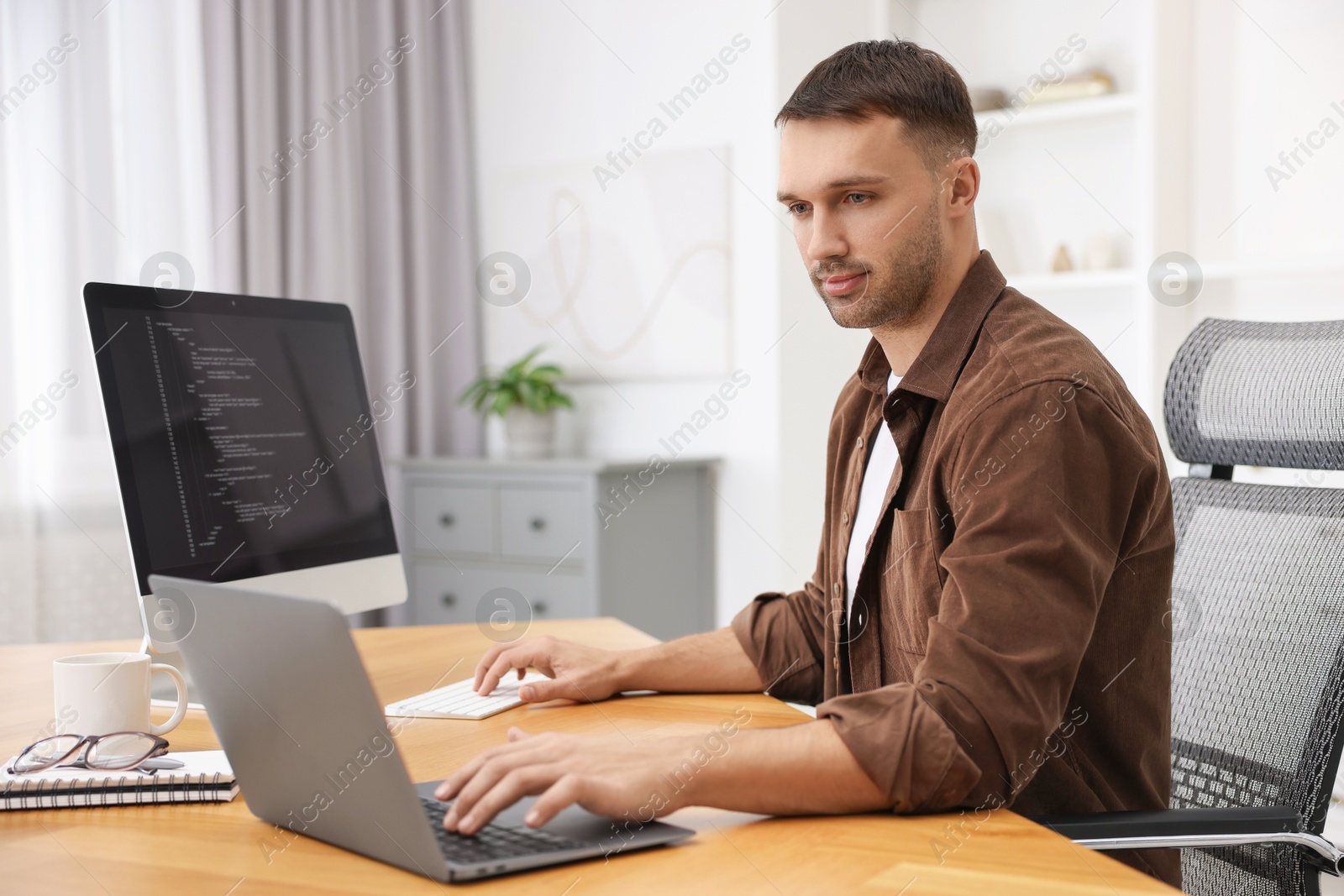 Photo of Programmer working on laptop and computer at wooden desk indoors