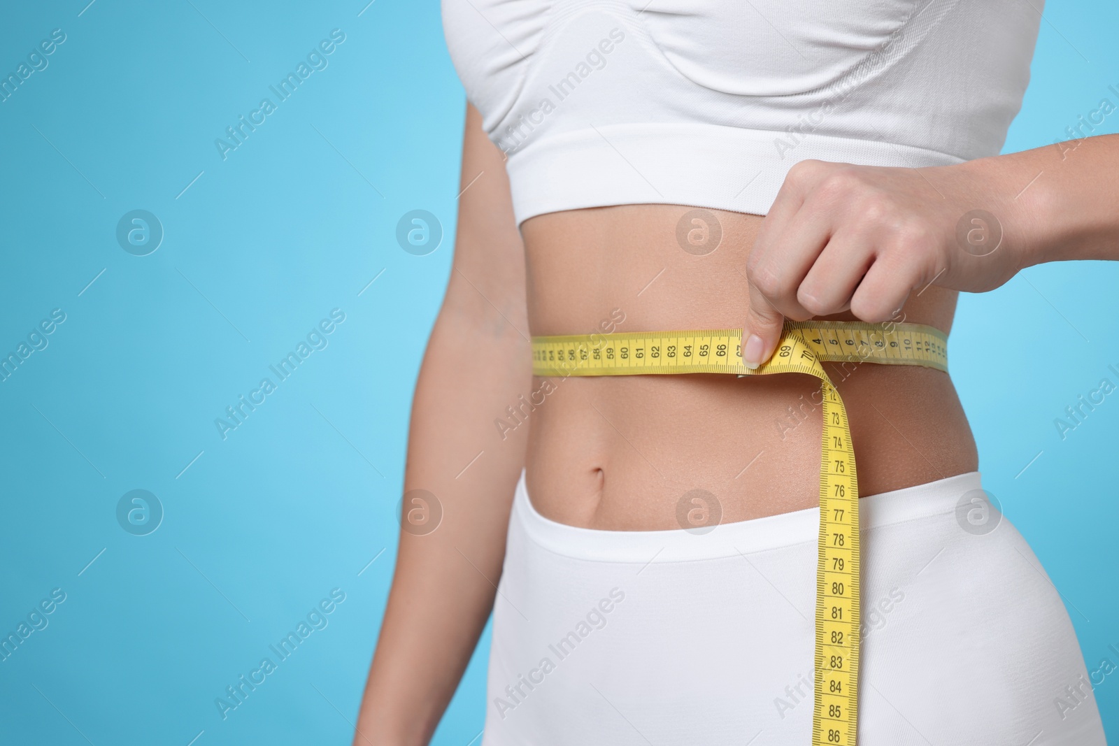 Photo of Weight loss. Woman measuring her slim waist with tape on light blue background, closeup. Space for text