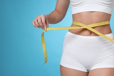 Photo of Weight loss. Woman measuring her slim waist with tape on light blue background, closeup. Space for text