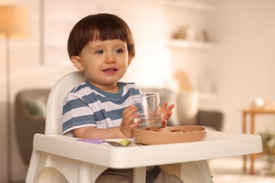 Photo of Cute little boy with glass of water in high chair at home