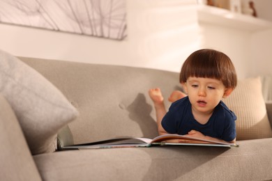 Photo of Cute little boy with book on sofa at home, space for text