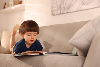 Photo of Cute little boy with book on sofa at home, space for text