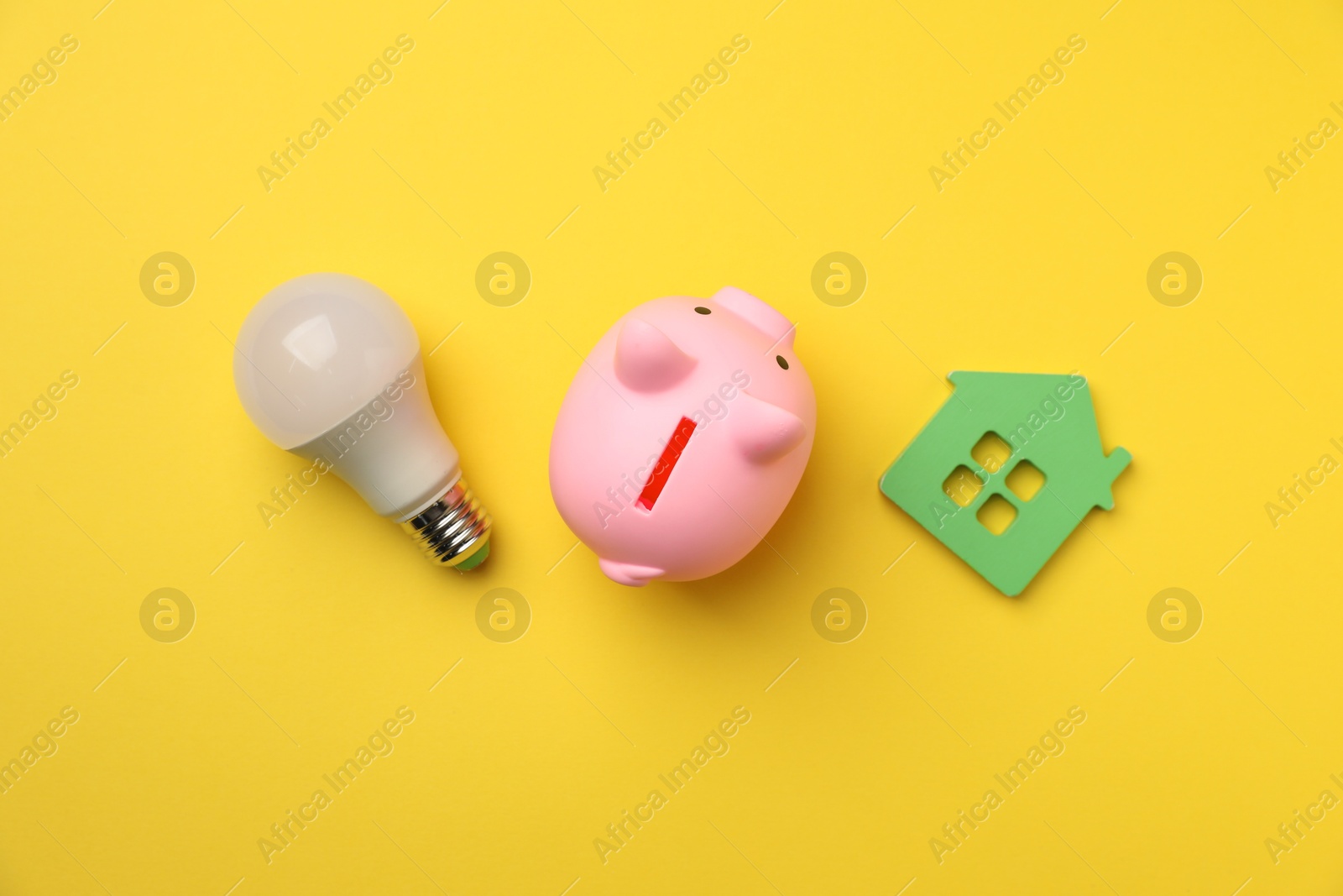 Photo of Piggy bank, light bulb and house model on yellow background, flat lay. Energy saving concept