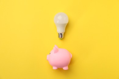 Photo of Light bulb and piggy bank on yellow background, flat lay. Energy saving concept