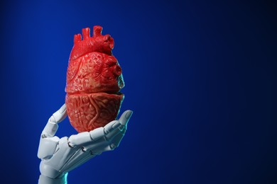 Photo of Artificial intelligence. Robot hand holding model of human heart on blue background, closeup. Space for text