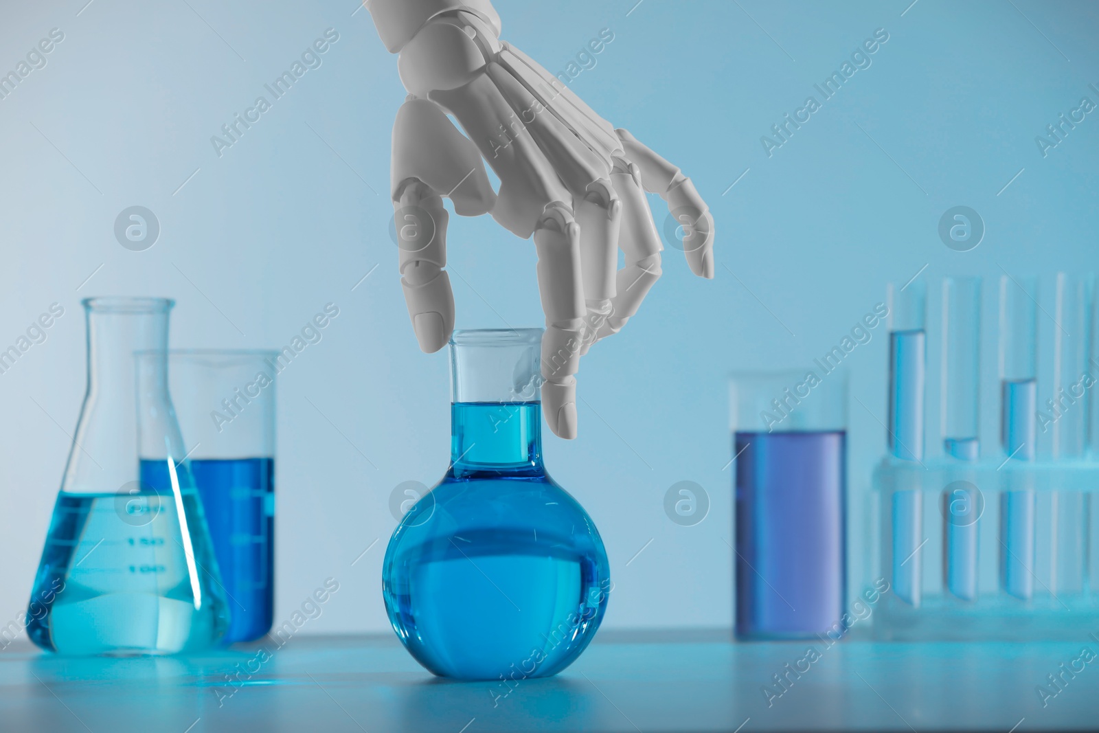 Photo of Artificial intelligence. Robot hand with flask and other laboratory glassware with liquids on table against light blue background