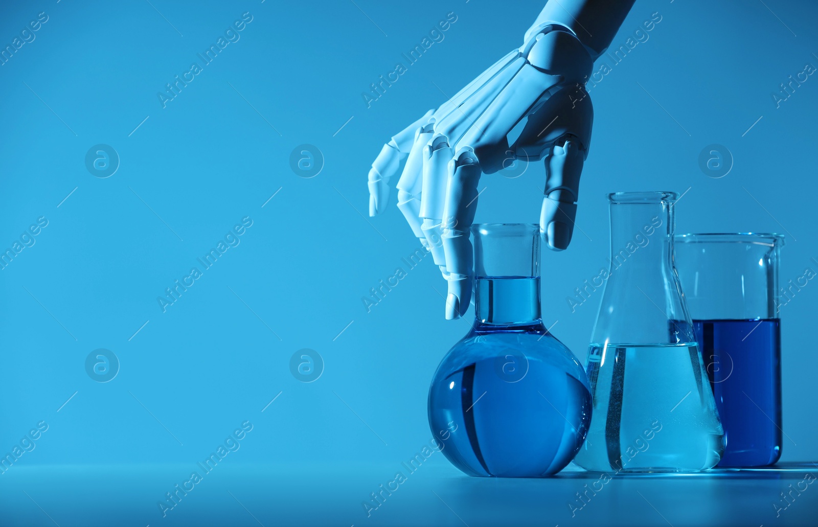 Photo of Artificial intelligence. Robot hand and laboratory glassware with liquids at table in blue light, space for text