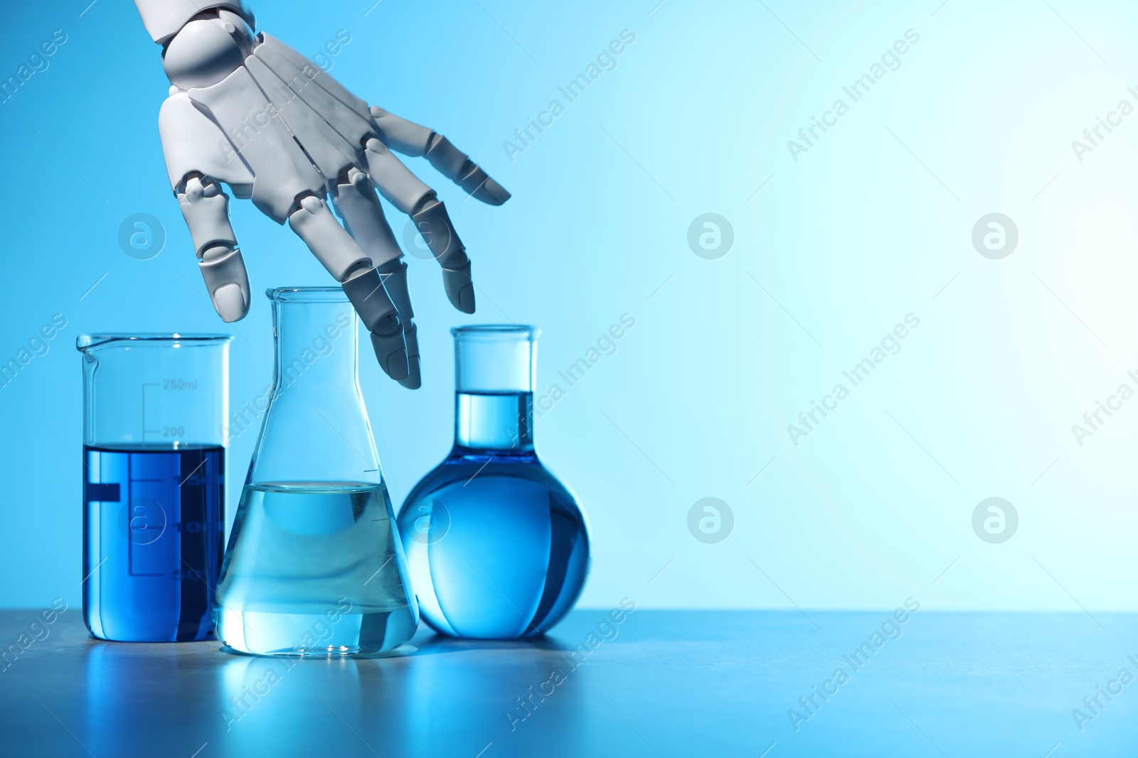 Photo of Artificial intelligence. Robot hand and laboratory glassware with liquids on table against light blue background, space for text