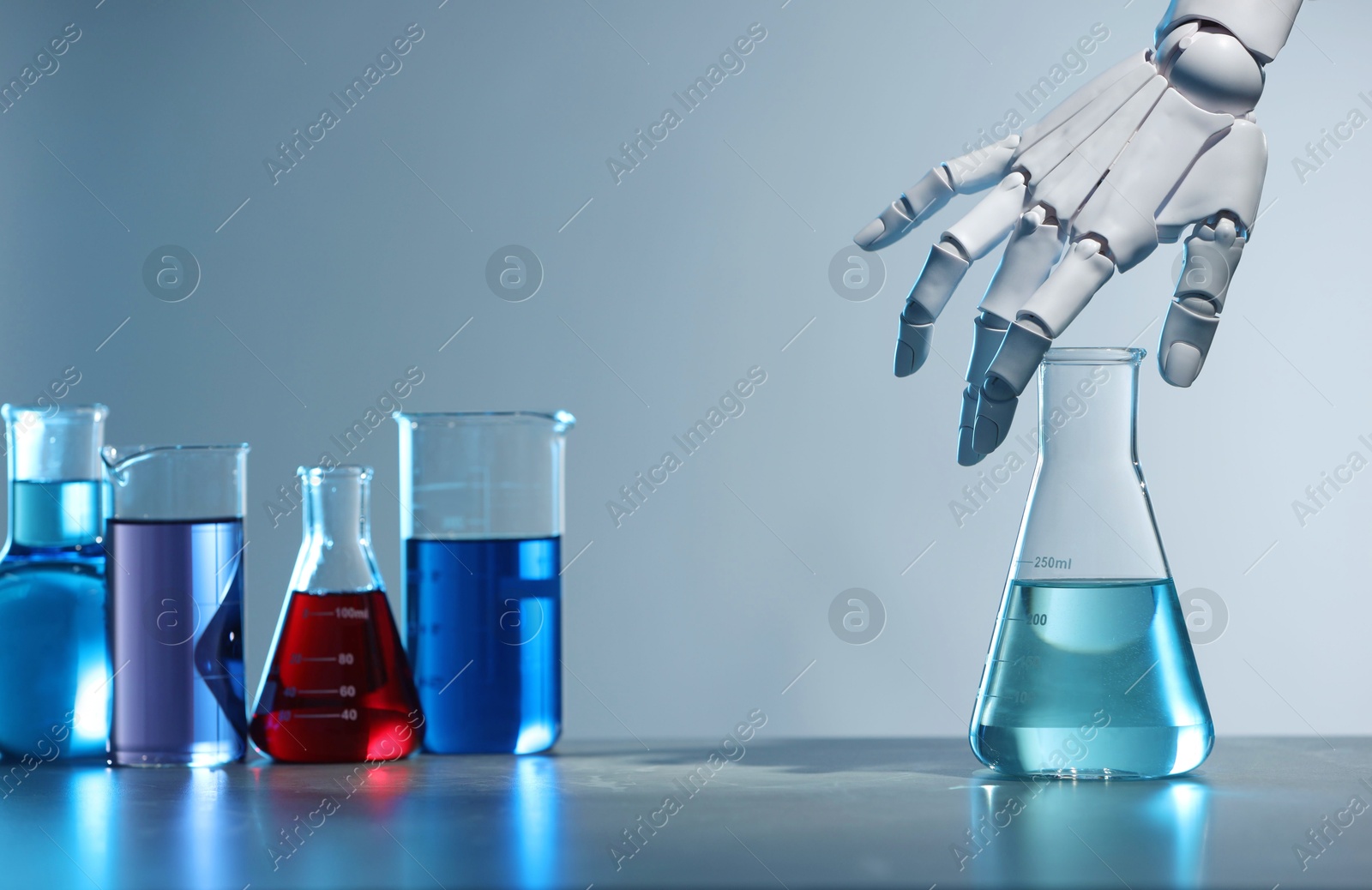 Photo of Artificial intelligence. Robot hand with flask and other laboratory glassware with liquids on table against light grey background