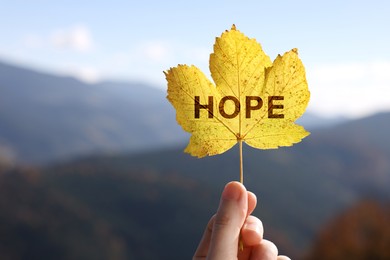 Image of Woman holding beautiful autumn leaf with word Hope in mountains, closeup