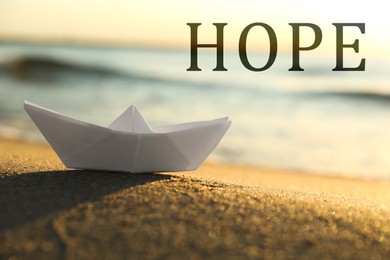 Image of Hope. White paper boat on sand near sea at sunrise, closeup