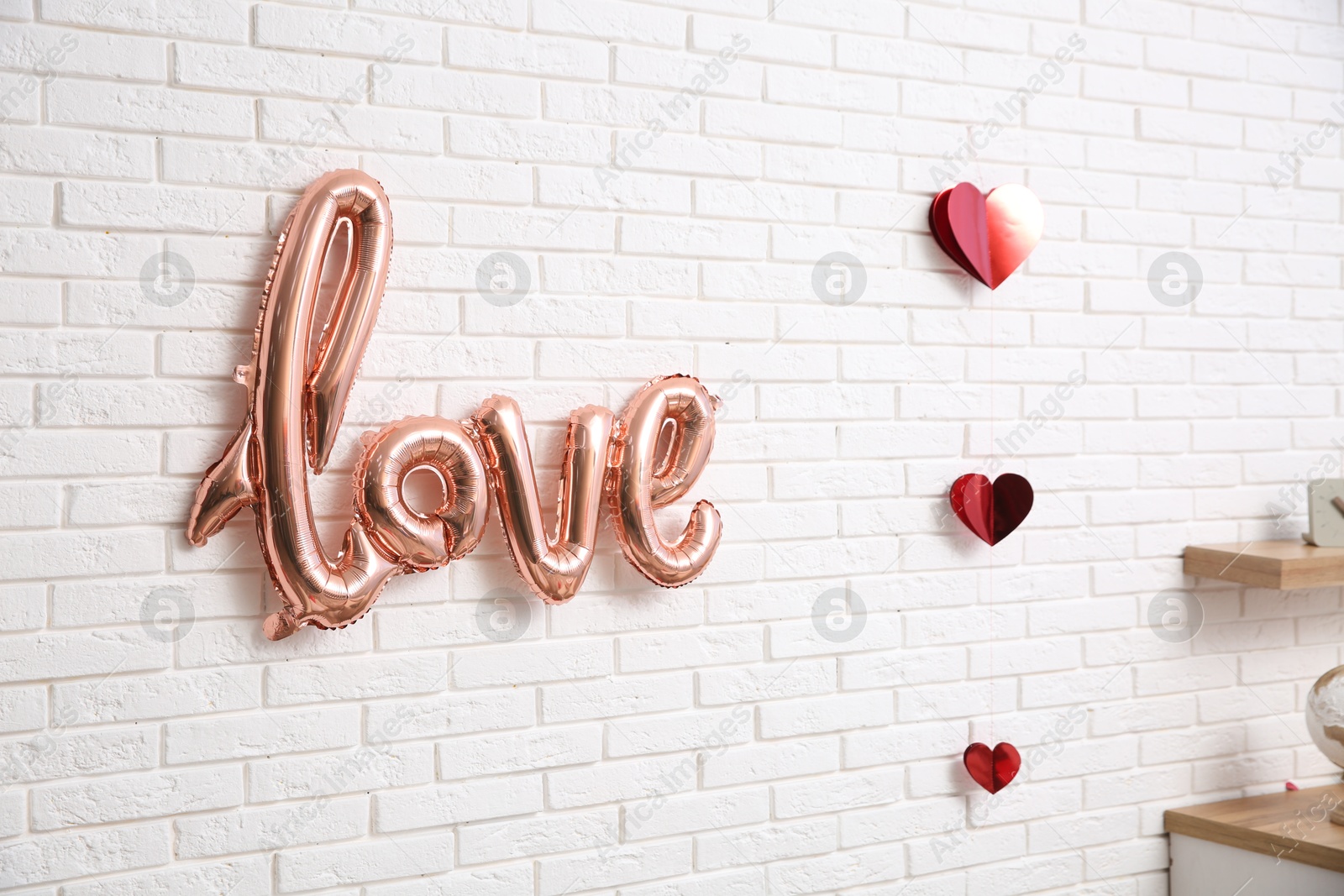 Photo of Pink foil balloon in shape of word Love and red hearts hanging on white brick wall indoors. Valentine Day celebration