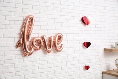 Photo of Pink foil balloon in shape of word Love and red hearts hanging on white brick wall indoors. Valentine Day celebration